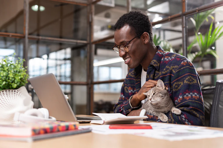 A man wearing glasses pets a cat while smiling into an open laptop.