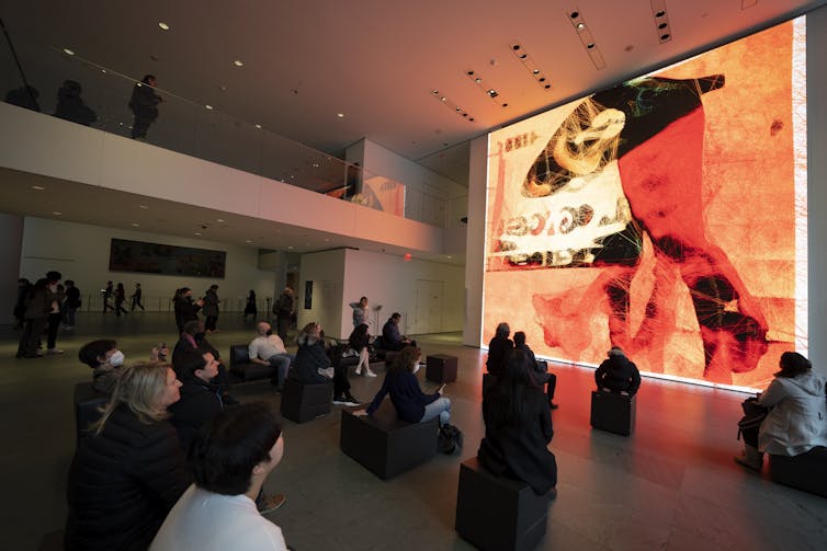 A group of people sit on the floor looking at a huge ceiling-high screen displaying an abstract artwork with an orange background and swatches of red, black, and ochre across it.