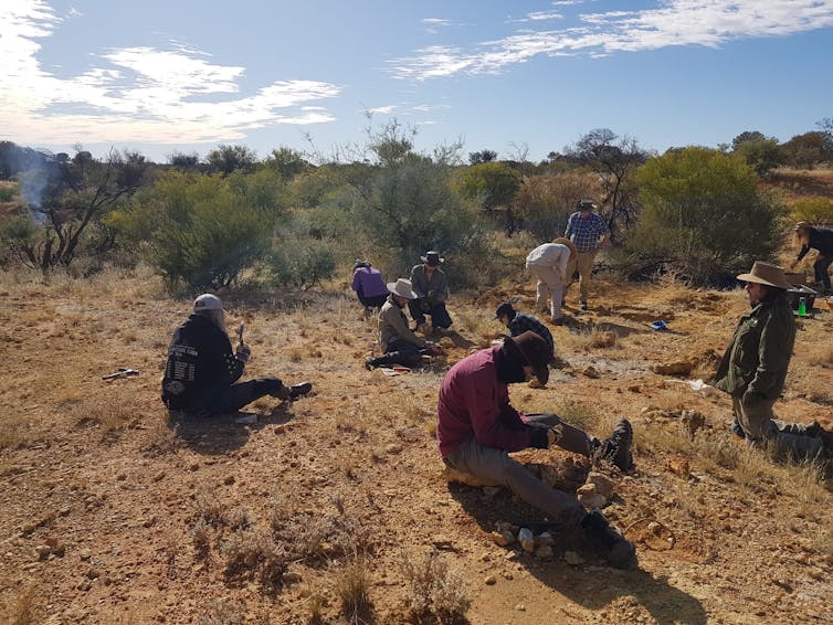A dry orange landscape with small shrubs and a group of people sifting through rocks in the foreground