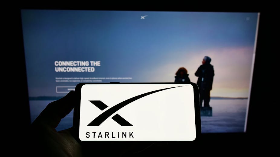 A hand holding a smartphone that features a black x and the word Starlink against a white background in front of a larger screen that reads Connecting the unconnected