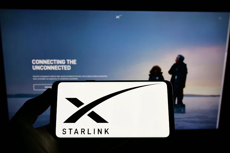 A hand holding a smartphone that features a black x and the word Starlink against a white background in front of a larger screen that reads Connecting the unconnected