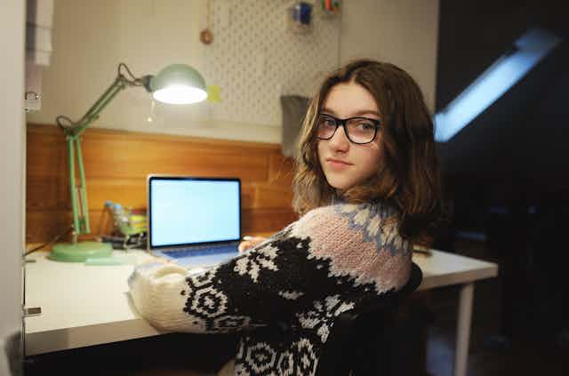 A teenager sitting at a desk with laptop and lamp, looking over her shoulder.