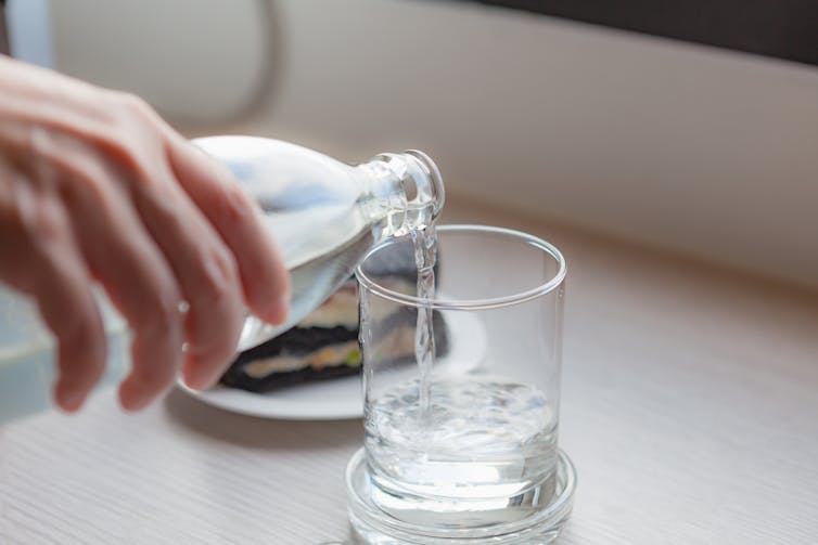 A hand, pouring a class of water, with a sandwich in the background.