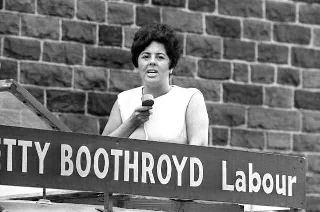 Betty Boothroyd pictured as a young woman holding a microphone standing behind a sign with her name on. 