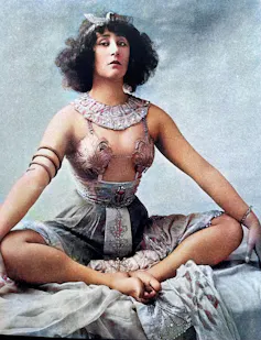 French author and actress Colette seated cross-legged, dressed in an exotic music hall costume from the 1920s.