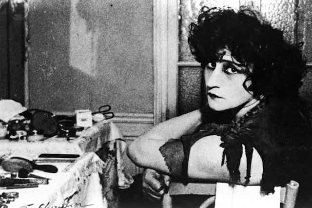 A clack and white photograph of French novelist Colette with very dark eye make-up sitting at a dresser.