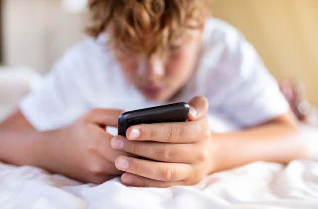 A boy laying on his bed looks at and holds his cell phone.