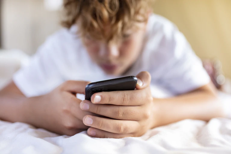 A boy laying on his bed looks at and holds his cell phone.