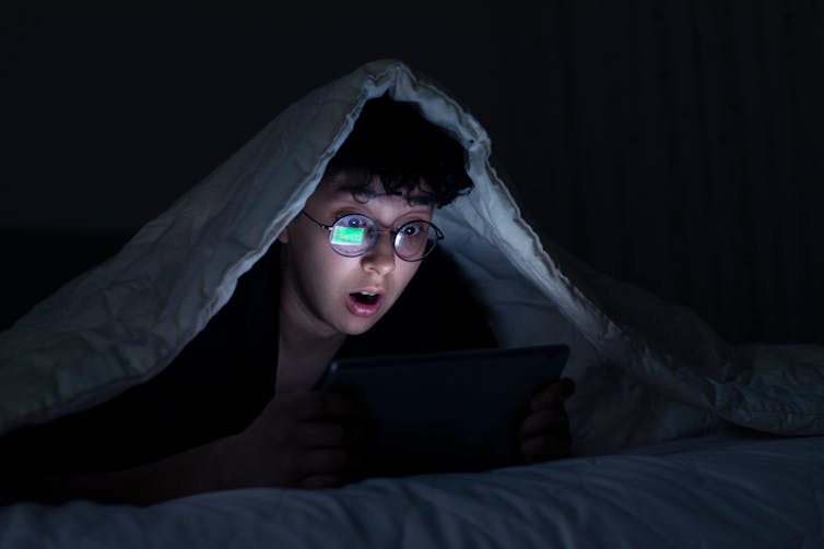 A girl with glasses is underneath a bedcover as the green glare of an iPhone illuminates her surprised and bespectacled face.