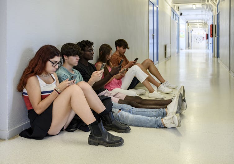 Five teenagers sit on the floor of a high school hallway with their backs to the wall as they look at their cell phones.