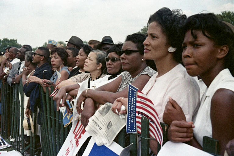 A line of people stand behind a flimsy green gate as they listen to a speech.