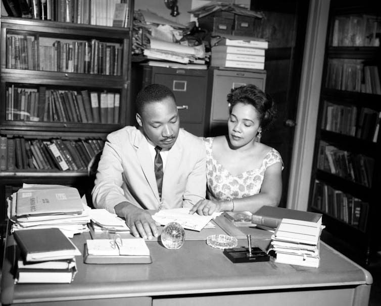 A man in a light-colored suit and a woman in short-sleeved dress look at a piece of paper together in a study.