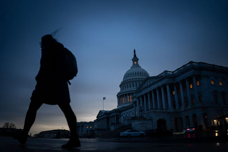 A person wearing a backpack walks past a large building with a white dome in a blueish dusk.