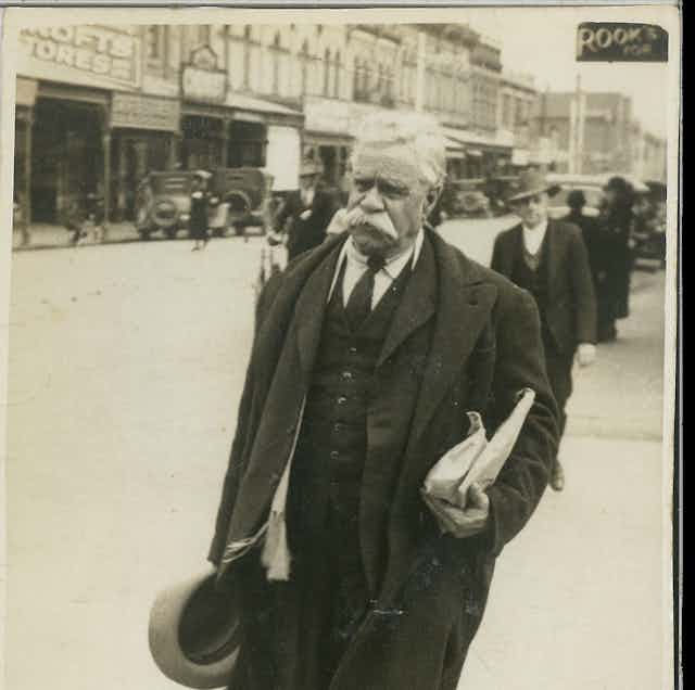 A black and white image of William Cooper, a senior Aboriginal man walking down a street. He holds something under his arm.