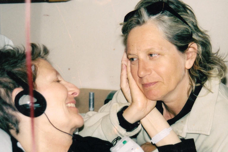 Two women in a hospital setting, one smiling from her bed and the other holding the sick woman's hand against her cheek with a look of love.