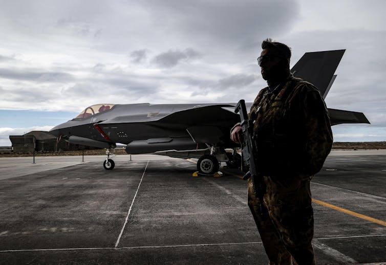 رجل يحمل بندقية يقف بجانب مقاتلة نفاثة على المدرج.