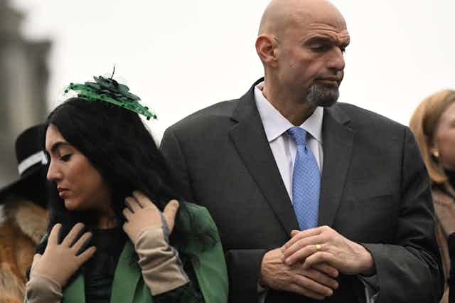 A dark-haired woman wearing gloves and a green hat standing with a large man in a dark suit and blue tie.