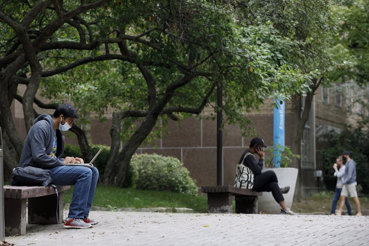 Students seen sitting on benches outdoors, one working on a laptop.