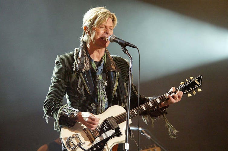 David Bowie with a white guitar on a stage.