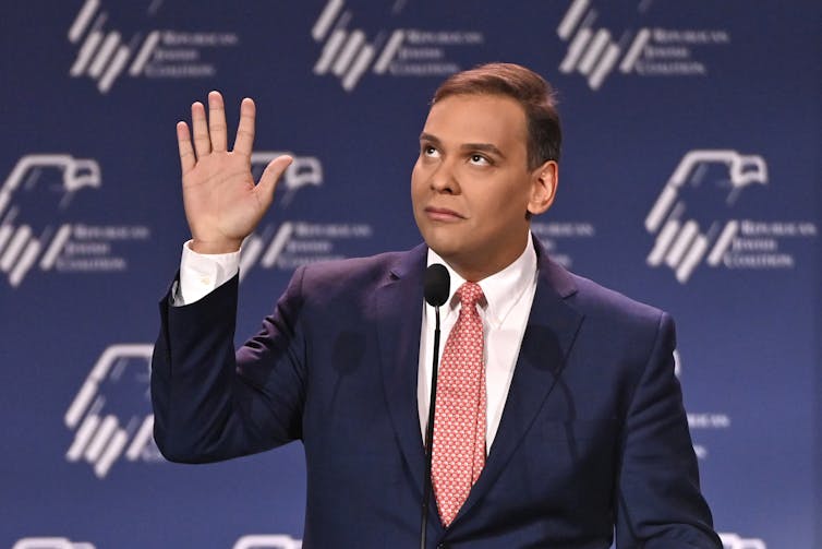 A man in a dark jacket, red tie and white shirt raising his right hand and looking upward.