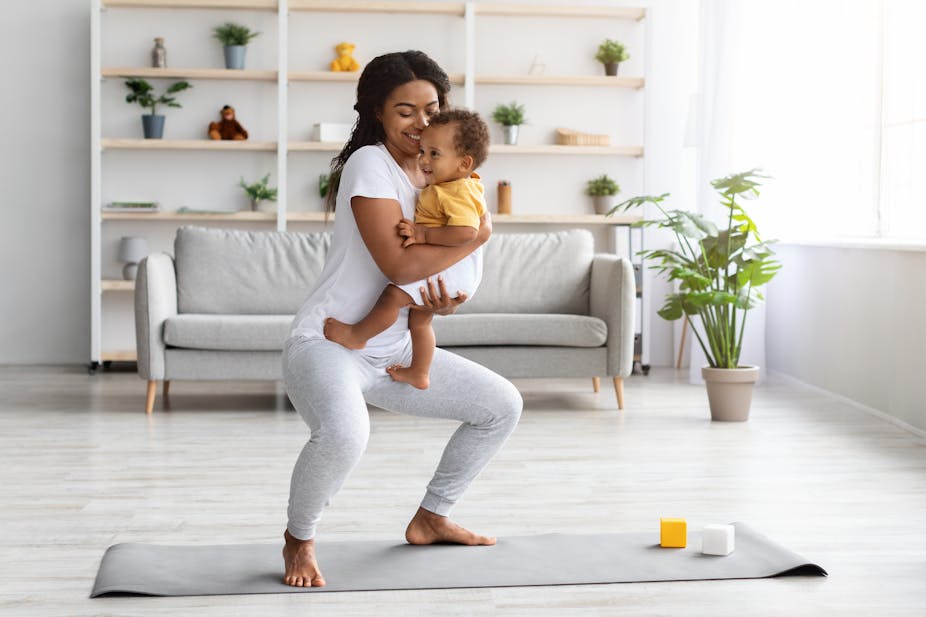 A young woman holds her baby while performing a squat in her living room.