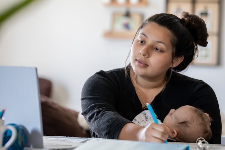 Young woman holds infant to chest while working in front of laptop