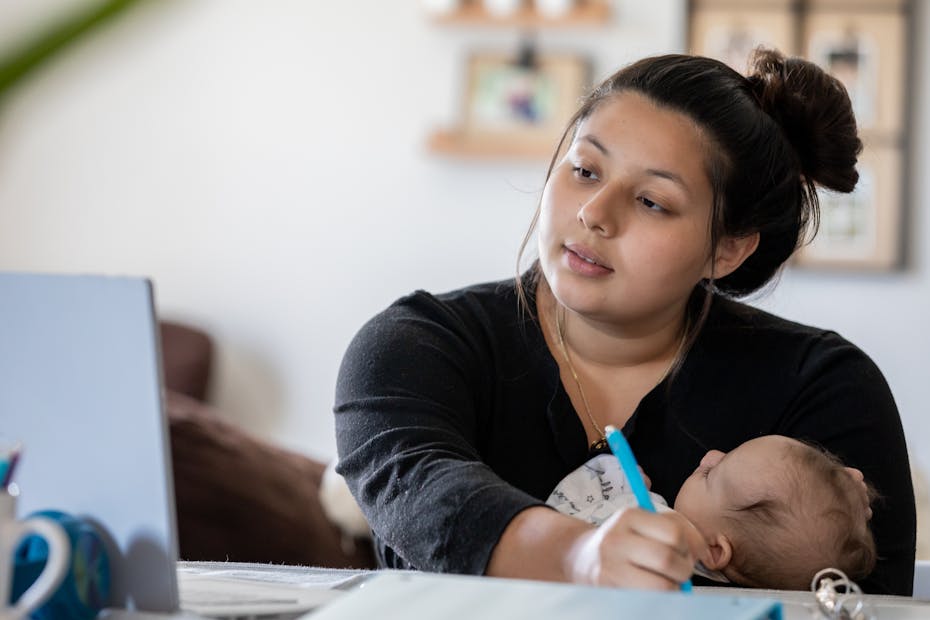 Young woman holds infant to chest while working in front of laptop
