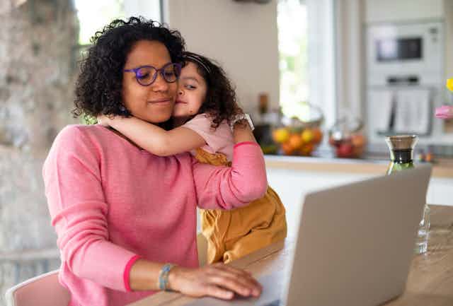 Mother wearing purple glasses hugs preschool-age daughter while working on laptop