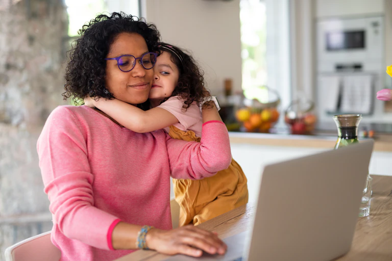 Mother wearing purple glasses hugs preschool-age daughter while working on laptop