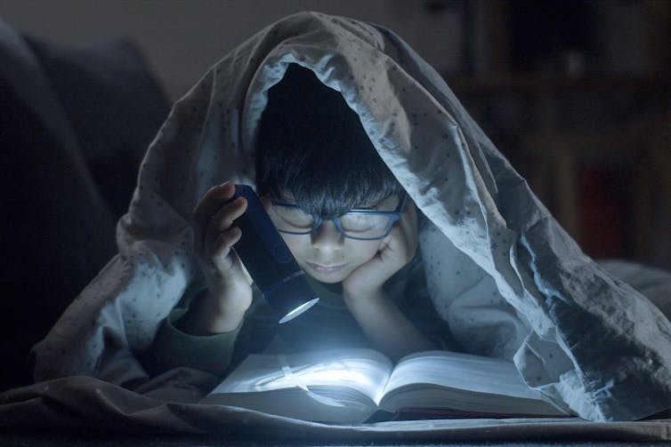 A child wearing glasses is reading a book with a flashlight under a blanket.