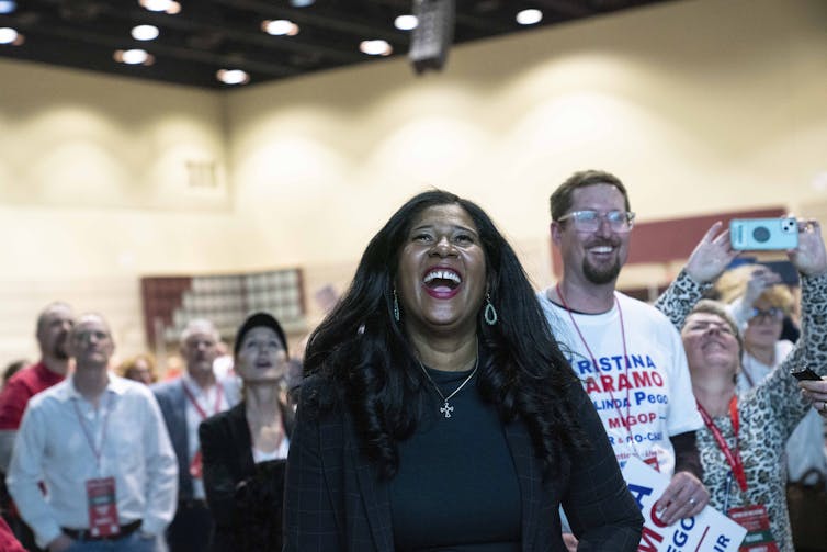 A smiling Black woman stands in front of a group of white men and women.