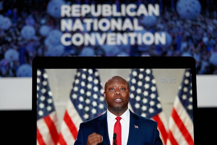 A Black man dressed in a dark suit standing in front of several American flags appears on a television screen