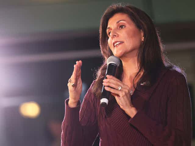 A brown-haired woman in a purple sweater gestures while holding a microphone.