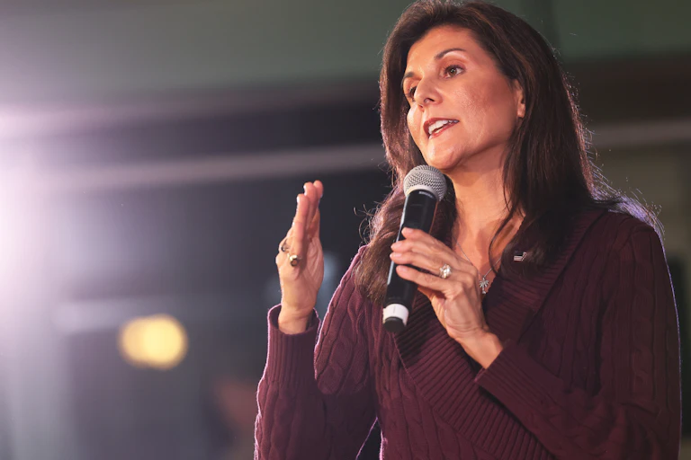 A brown-haired woman in a purple sweater gestures while holding a microphone.