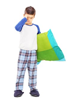 A boy in pyjamas rubbing his eye, carrying a pillow