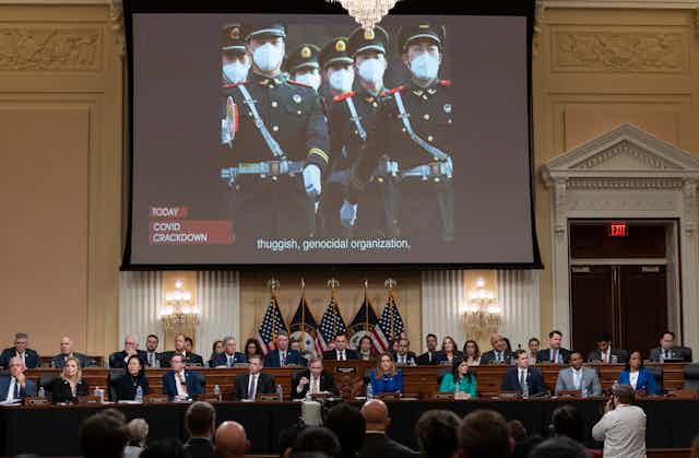A group of politicians on two rows of bench sit below a t giant screen showing Chinese troops in uniform.