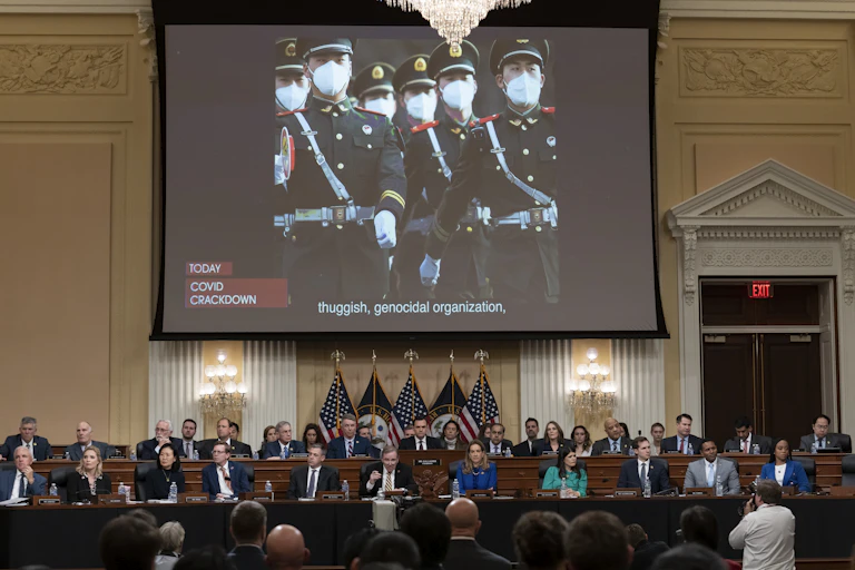 A group of politicians on two rows of bench sit below a t giant screen showing Chinese troops in uniform.