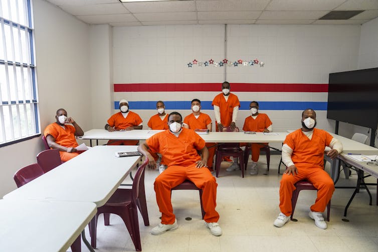 Men in bright orange outfits and face masks sit in what looks like an empty classroom with white bars on the windows.