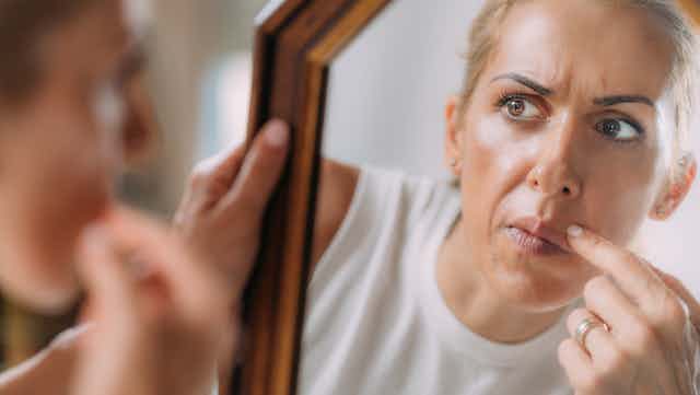 An adult woman, with blond hair, brown eyes, and a concerned look, examines her facial features in a hand-held mirror.