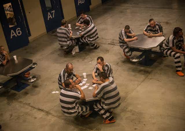 People wearing white and black striped outfits sit around tables together in a barren room. 