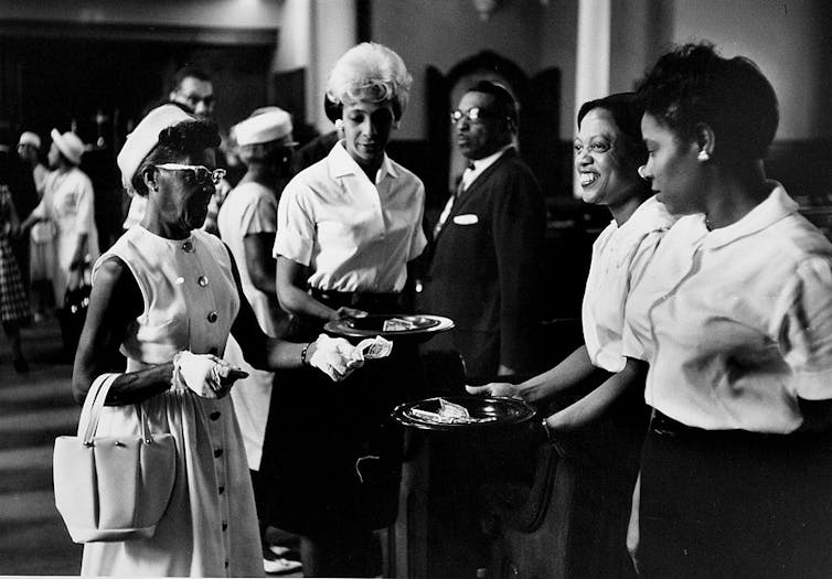 A black and white photo shows several formally dressed women putting money in a church collection plate.