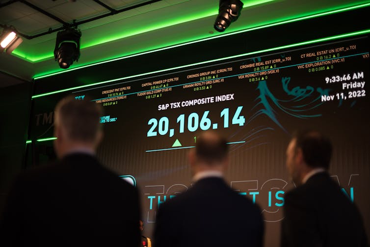 A crowd of people in suits look at a large screen that says 'S&P TSX composite index' on it