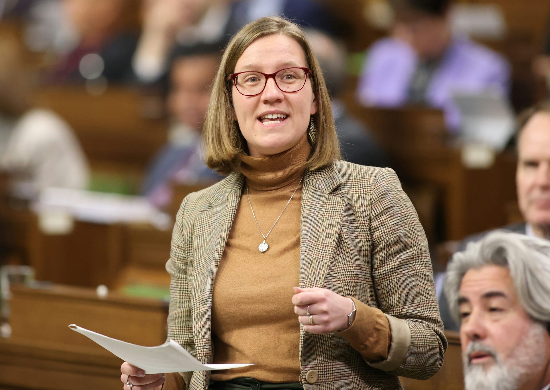 A women with shoulder-length blonde hair and glasses, wearing a blazer and turtleneck, gestures while speaking in the House of Commons