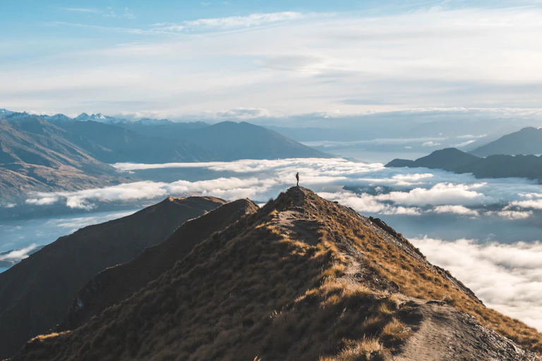 A lone figure standing on a hilltop with mountains and clouds in the background