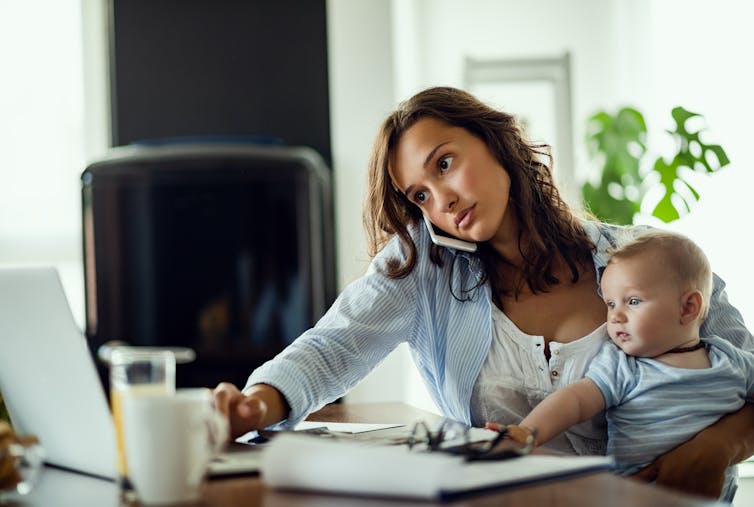 A woman holds a baby while typing on the computer and talking on the phone.