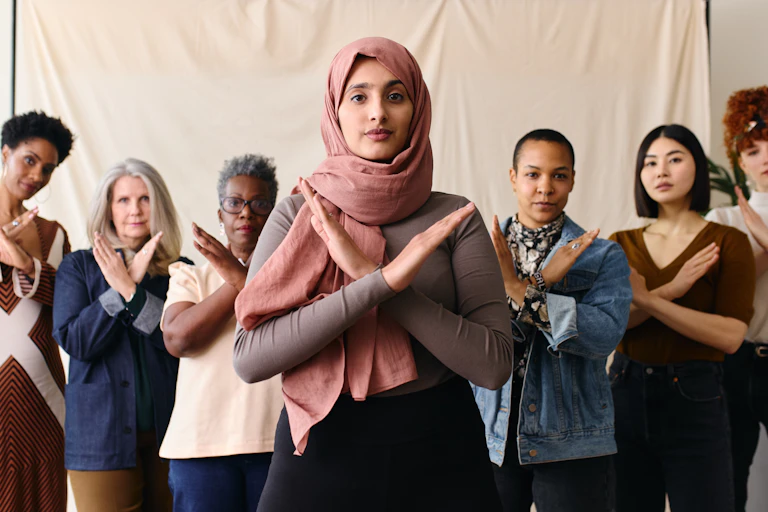 A group of women from varying ethnical and racial backgrounds standing in a row with their arms crossed in front of their chests in an 'X' sign