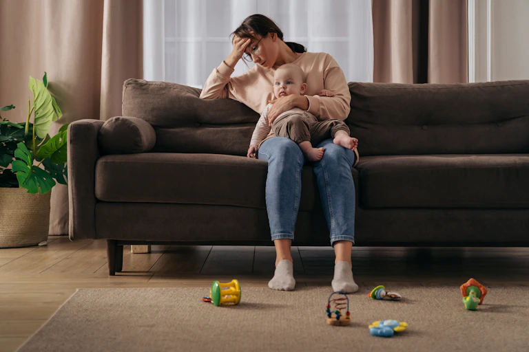 A woman sits on a couch while holding a baby. She is resting her head against her hand with a tired look on her face.