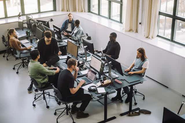 eight people work on computers in an open office with large windows