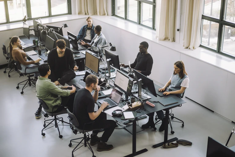 eight people work on computers in an open office with large windows