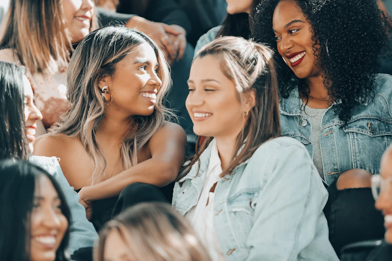 women seated in a large crowd, two looking at each other and smiling, one looking thoughtful